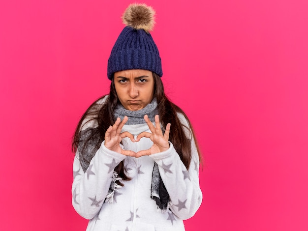 Triest ziek jong meisje dat de winterhoed met sjaal draagt die hartgebaar toont dat op roze wordt geïsoleerd