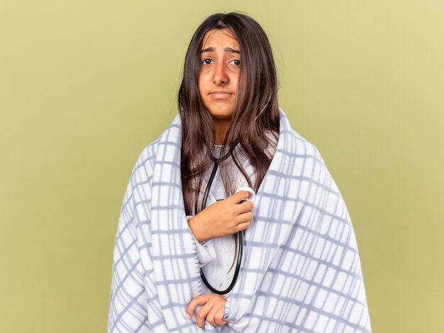 Triest ziek meisje gewikkeld in plaid dragen en luisteren naar haar eigen hartslag met een stethoscoop