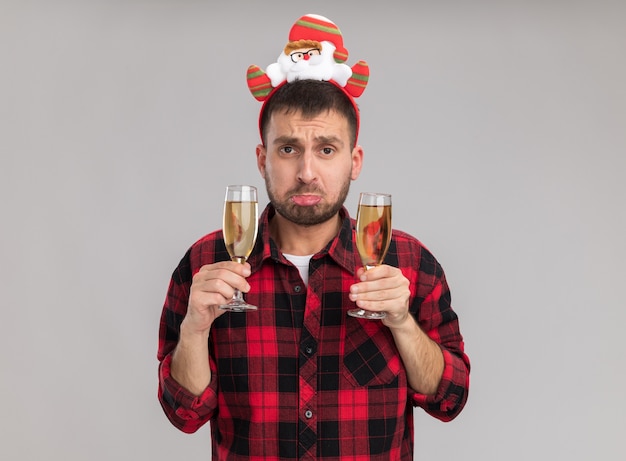 Trieste jonge blanke man met kerst hoofdband met twee glazen champagne kijken camera geïsoleerd op een witte achtergrond met kopie ruimte