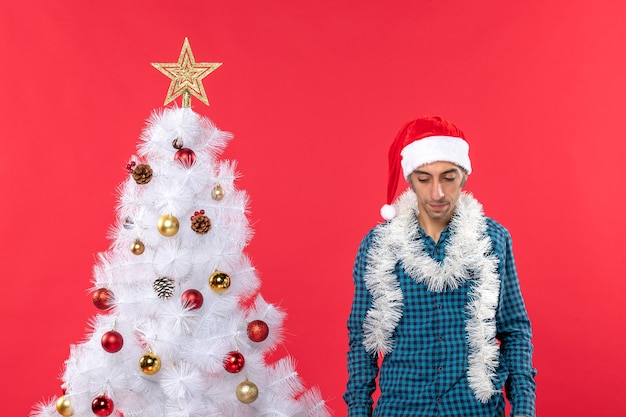 Trieste jonge man met kerstman hoed in een blauw gestript shirt en neerkijkt staande in de buurt van kerstboom op rood