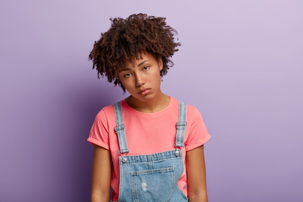 Trieste jonge vrouw heeft afro-kapsel, apathie in de ogen, kantelt het hoofd, staat niet te popelen om iets te doen na hard werken, voelt zich overwerkt, draagt een casual t-shirt, spijkerbroek, ziet er ongelukkig uit recht in de camera