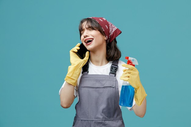 Trieste jonge vrouwelijke schoonmaker met uniforme bandana en rubberen handschoenen met reinigingsmiddel pratend aan de telefoon met gesloten ogen geïsoleerd op blauwe achtergrond