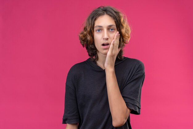 Trieste man met lang haar in zwart t-shirt legde zijn hand op pijnlijke tand op roze muur