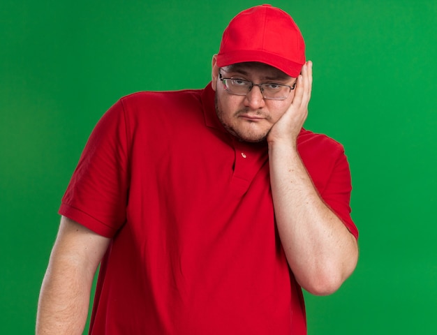 trieste overgewicht jonge bezorger in optische bril legt hand op gezicht geïsoleerd op groene muur met kopieerruimte
