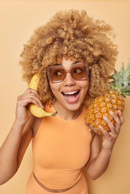 Tropisch fruitseizoen Vrolijke vrouw met blond krullend haar dwaast rond doet alsof telefoongesprek banaan in de buurt van oor in de ene hand verse ananas in andere draagt sportkleding en zonnebril