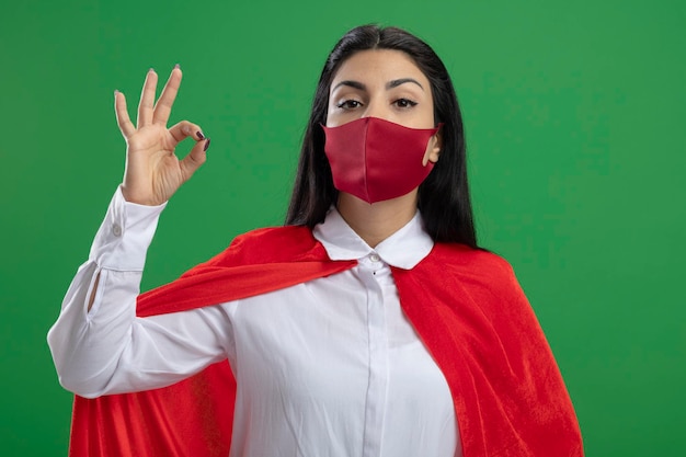 Trotse jonge superherovrouw die masker draagt dat het is in orde teken kijkt naar voorzijde geïsoleerd op groene muur