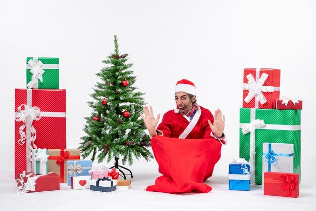 Trotse jonge volwassene verkleed als kerstman met geschenken en versierde kerstboom zittend op de grond op witte achtergrond