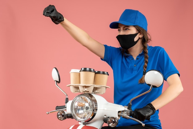 Trotse vrouwelijke koerier met een zwart medisch masker en handschoenen die bestellingen afleveren op perzikachtergrond