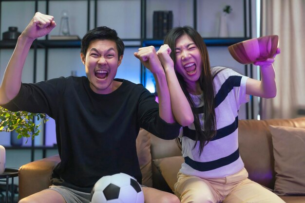 Trouwen met volwassen aziatische tv-kijken thuis aziatisch paar juichen sportgames competitie samen met lach glimlach overwinning op sofa bank bij woonkamer thuis isolatie-activiteit