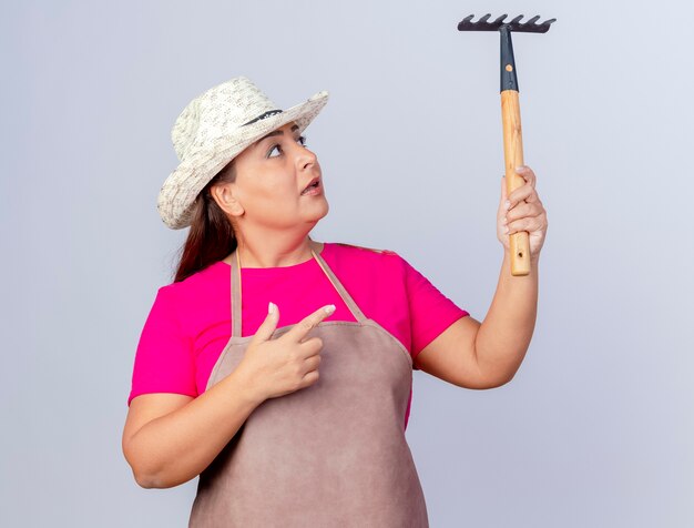Gratis foto tuinmanvrouw van middelbare leeftijd in schort en hoed die een mat vasthoudt en ernaar wijst met de wijsvinger