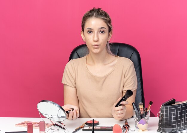 Tuitende lippen jong mooi meisje zit aan tafel met make-up tools met poeder borstel geïsoleerd op roze achtergrond