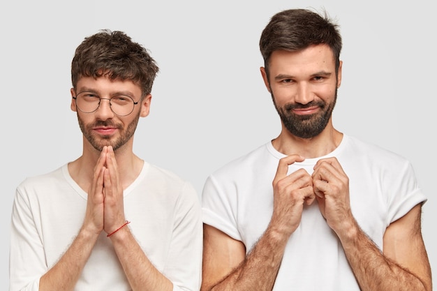 Twee aantrekkelijke ongeschoren jonge mannen hebben een intrigerende uitstraling, houden elkaars handen, gekleed in vrijetijdskleding, geïsoleerd over een witte muur. knappe hipsters poseren binnen