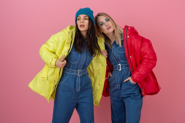 Gratis foto twee aantrekkelijke stijlvolle vrouwen poseren op roze muur in kleurrijke winter donsjack van rode en gele kleur, warme kleding modetrend