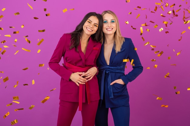 Twee aantrekkelijke vrouwen vieren nieuwjaar op violette muur in stijlvolle kleurrijke avondpakken van paarse en blauwe kleur, vrienden samen plezier hebben, modetrend, gouden confetti feeststemming