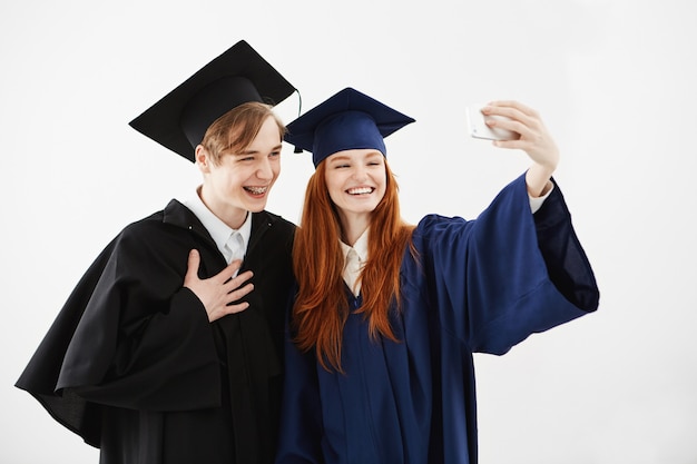 Twee afgestudeerde vrienden in petten en mantels lachen en maken selfie voordat ze hun magister-diploma of bachelor of arts of een andere academische graad ontvangen. Studie concept.