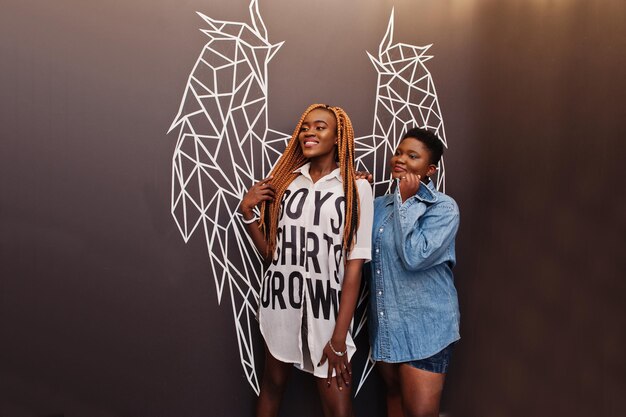 Twee Afrikaanse dames in stijlvolle vrijetijdskleding poseren tegen de muur van engelenvleugels