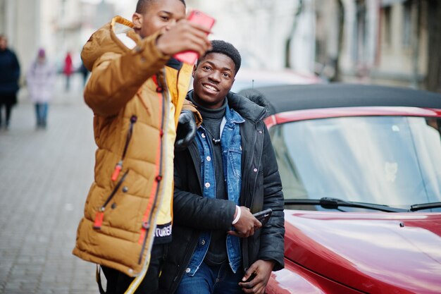 Twee Afrikaanse mannelijke vrienden die zich dichtbij rode Amerikaanse convertibele auto bevinden en selfie op telefoon maken