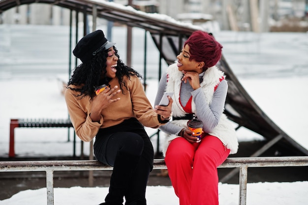 Twee Afro-Amerikaanse dames poseerden op een winterdag tegen een besneeuwde achtergrond met kopjes koffie en kijken op de telefoon