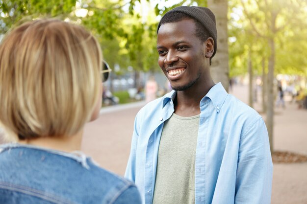 Twee beste vrienden ontmoeten elkaar op straat: een man met een donkere huidskleur in trendy hoed en overhemd, breed glimlachend terwijl ze een gesprek heeft met zijn vriendin, blij haar per ongeluk te ontmoeten in het groene park