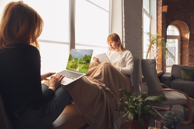 Twee blanke vrouwelijke modellen thuis in de buurt van venster met behulp van laptop en tablet.