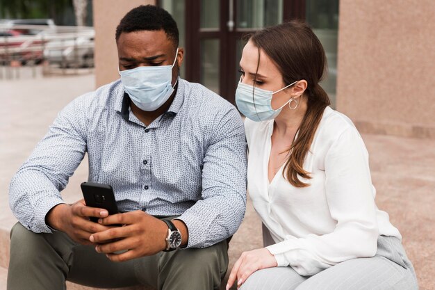 Twee collega's kijken naar smartphone buitenshuis tijdens pandemie met gezichtsmaskers