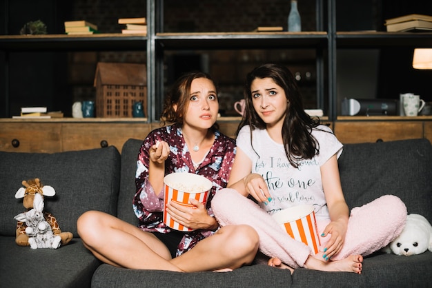 Twee doen schrikken vrouwen die op bank met popcorn zitten die op televisie letten