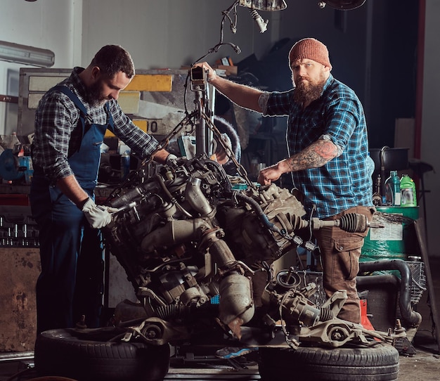 Gratis foto twee gebaarde werktuigkundigenspecialist repareert de motor van een auto die op de hydraulische lift in de garage wordt opgeheven. servicestation.