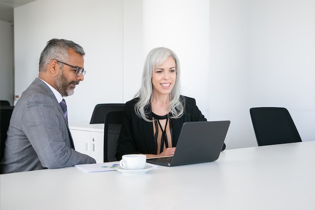 Twee gelukkige collega's met behulp van laptop voor videogesprek, zittend aan tafel met een kopje koffie, beeldscherm kijken en praten. Online communicatieconcept