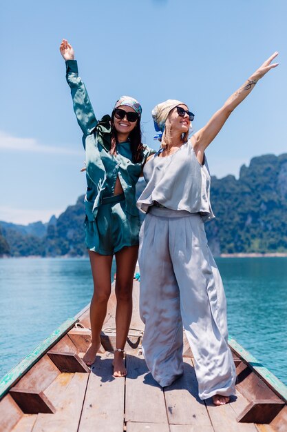 Twee gelukkige de toeristenvrienden van de vrouwenblogger in zijden kostuum en sjaal en zonnebril op vakantiereis rond thailand op Aziatische boot, nationaal park Khao Sok.