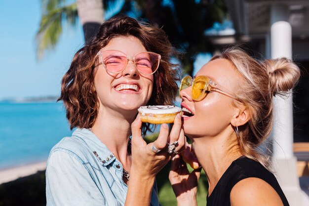 Twee gelukkige fit vrouw in roze en gele zonnebril lachend plezier lachen met donuts, buiten