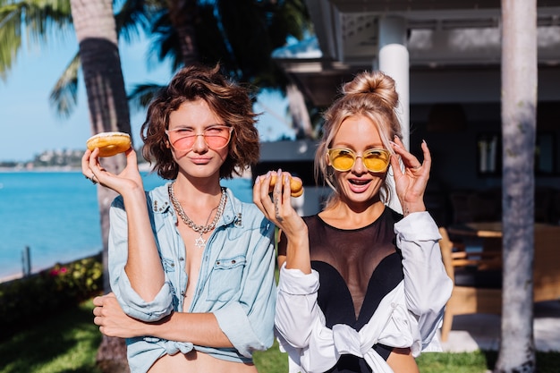 Twee gelukkige fit vrouw in roze en gele zonnebril lachend plezier lachen met donuts, buiten