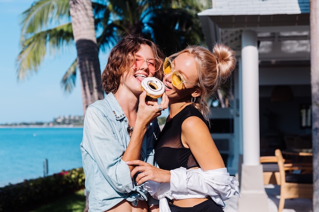 Twee gelukkige fit vrouw in roze en gele zonnebril lachend plezier lachen met donuts, buiten