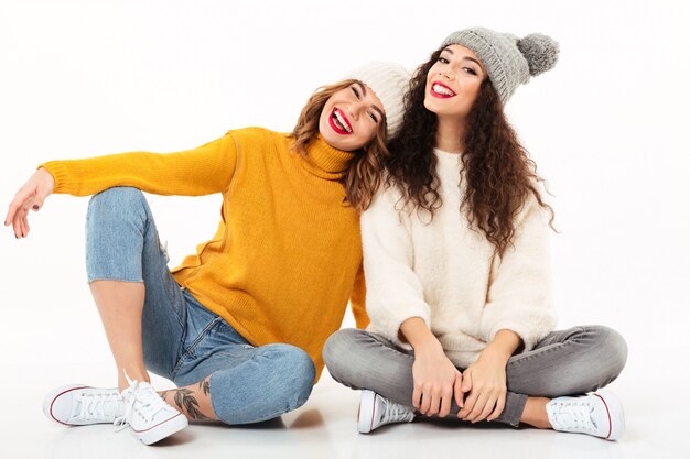 Twee gelukkige meisjes die in sweaters en hoeden op de vloer samen over witte muur zitten