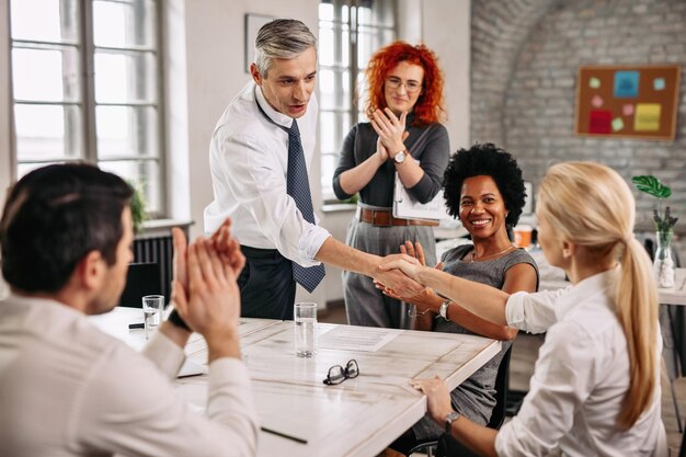 Twee gelukkige zakenmensen schudden elkaar de hand tijdens een vergadering terwijl andere collega's hen applaudisseren op kantoor