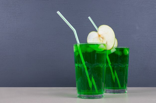 Twee glazen groen sap met rietjes op blauwe tafel.