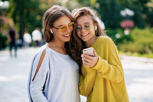 Twee grappige lachende zusters selfie maken op smaptphone en muziek luisteren, poseren op straat, vakantiestemming, gek positief gevoel, zomer lichte kleding zonnebril.