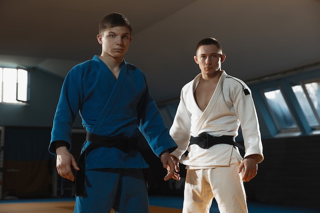 Twee jonge blanke judo-jagers in witte en blauwe kimono met zwarte gordels die zelfverzekerd in de sportschool poseren, sterk en gezond. Vechtvaardigheden in vechtsporten oefenen. Overwinnen, doel bereiken.