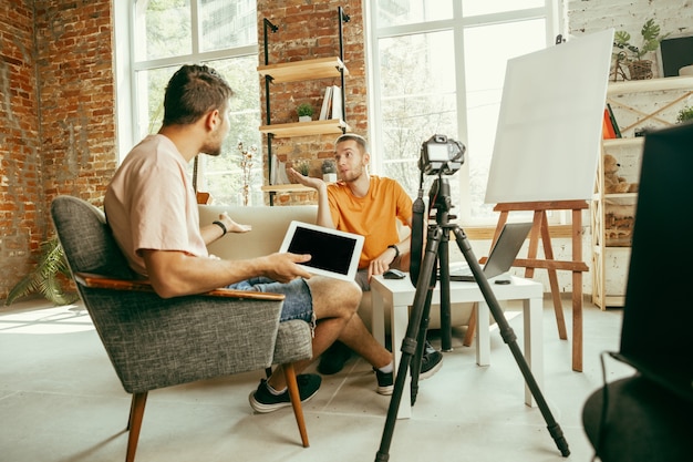 Twee jonge blanke mannelijke bloggers in vrijetijdskleding met professionele apparatuur of camera opname video-interview thuis