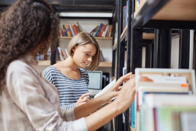 Twee jonge charmante studentenmeisjes die zich dichtbij boekenplank bevinden, informatie zoeken voor groepsproject in bibliotheek, sprekend over punten van teamwerk.