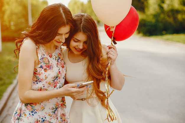 twee jonge en heldere meisjes brengen hun tijd door in het zomerpark met ballonnen en telefoon