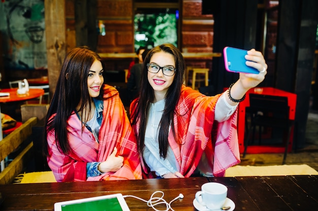 Twee jonge en mooie meisjes zitten aan tafel en doen selfie in het café