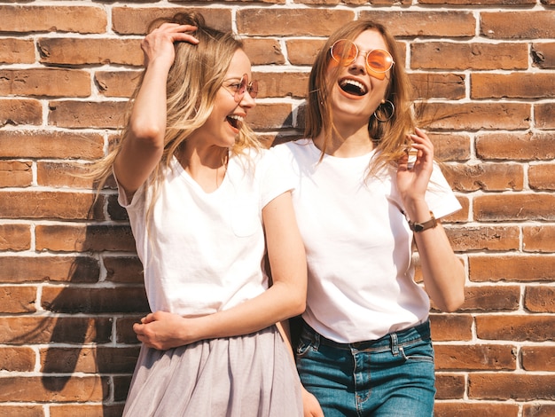 Twee jonge mooie blonde glimlachende hipster meisjes in trendy zomer wit t-shirt kleding. . Positieve modellen die pret in zonnebril hebben