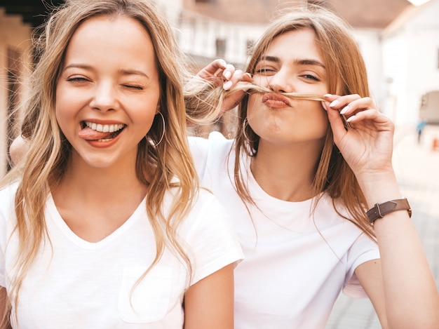 Twee jonge mooie blonde glimlachende hipster meisjes in trendy zomer wit t-shirt kleding. Positieve modellen met plezier. Snor maken met haar en tong laten zien