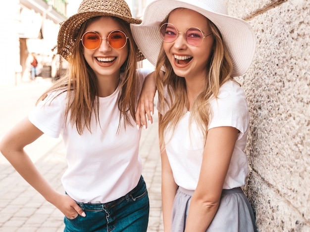 Twee jonge mooie blonde glimlachende hipster meisjes in trendy zomer wit t-shirt kleding. vrouwen die zich in de straat dichtbij muur stellen.