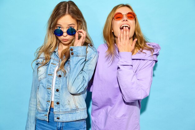 Twee jonge mooie blonde glimlachende hipster vrouwen in trendy de zomerkleren. Sexy onbezorgde vrouwen die dichtbij blauwe muur in zonnebril stellen. Positieve modellen worden gek