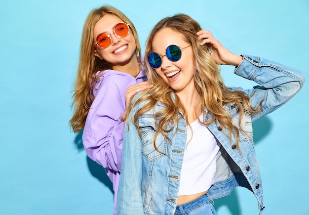 Twee jonge mooie blonde glimlachende hipster vrouwen in trendy de zomerkleren. Sexy onbezorgde vrouwen die dichtbij blauwe muur in zonnebril stellen. Positieve modellen worden gek