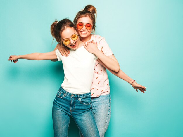 Twee jonge mooie blonde lachende hipster meisjes in trendy zomer hipster jeans kleding. sexy onbezorgde vrouwen die dichtbij blauwe muur stellen. trendy en positieve modellen die plezier hebben in zonnebrillen