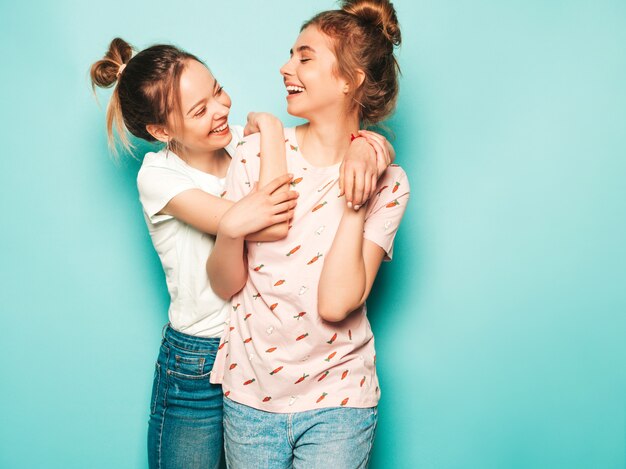 Twee jonge mooie blonde lachende hipster meisjes in trendy zomer hipster jeans kleding. Sexy onbezorgde vrouwen die dichtbij blauwe muur stellen. Trendy en positieve modellen die plezier hebben