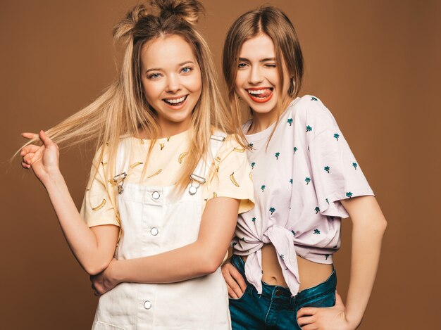 Twee jonge mooie glimlachende blonde hipstermeisjes in kleren van de trendy de zomer kleurrijke T-shirt. Sexy onbezorgde vrouwen die op beige achtergrond stellen. Positieve modellen hebben plezier