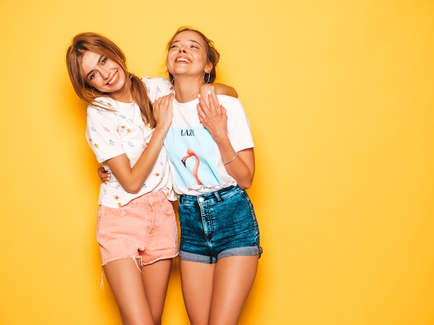 Twee jonge mooie glimlachende hipster meisjes in trendy zomerkleren. Sexy onbezorgde vrouwen die dichtbij gele muur stellen. Positieve modellen worden gek en hebben plezier